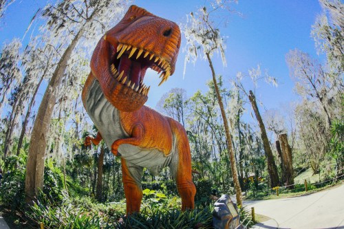 A large, orange dinosaur statue stands among trees draped with Spanish moss under a clear blue sky.