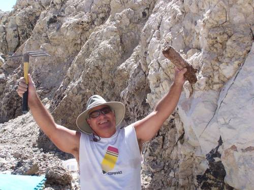 A smiling man in a hat holds a rock and a tool, standing in front of a rocky cliff.