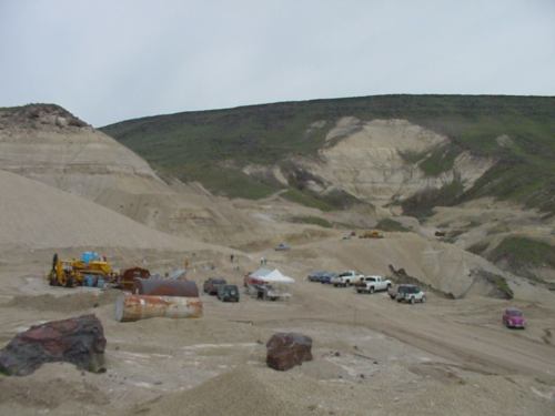 A gravel pit with machinery and vehicles on a sandy landscape, surrounded by hills under a cloudy sky.