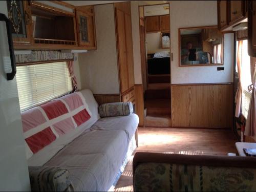 Interior of a cozy RV featuring a sofa, wooden cabinets, and a mirror, with a glimpse of a bedroom in the background.