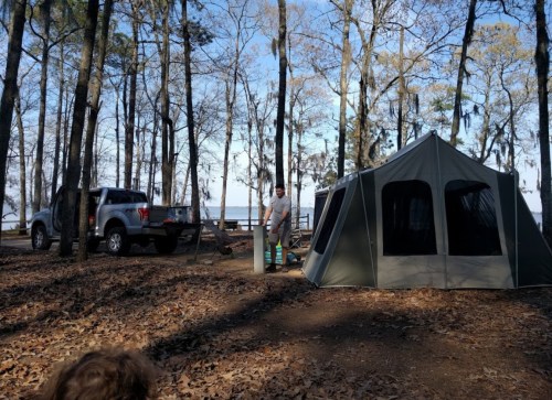 A campsite in the woods with a tent, a truck, and a person preparing near a lake surrounded by trees.