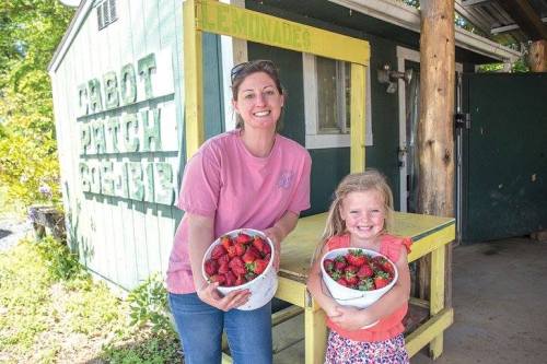 A woman and a girl hold bowls of strawberries outside a green building, smiling happily.