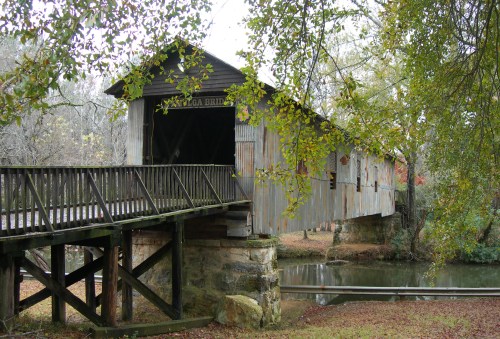 A rustic covered bridge with a wooden walkway, surrounded by trees and reflecting in a calm river.