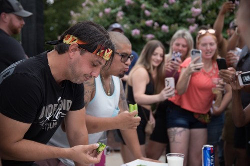 Two men compete in a food challenge while a crowd of onlookers records the event on their phones.