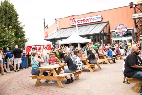 A lively outdoor scene with people dining at picnic tables near a venue called Victory, surrounded by a festive atmosphere.