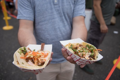 A person holds two tacos in paper trays, filled with colorful toppings and garnishes, at an outdoor food event.