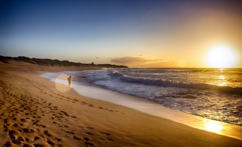 A person walks along a sandy beach at sunset, with gentle waves lapping at the shore and footprints in the sand.