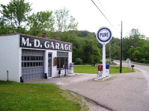 A vintage gas station with "M.D. Garage" sign, old fuel pumps, and a "Pure" gas sign, surrounded by greenery.