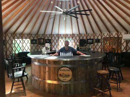 A person stands behind a circular wooden bar in a yurt, with a spacious interior and large ceiling fan above.