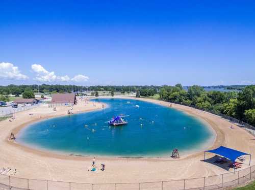 Aerial view of a sandy beach area with a blue lagoon, people swimming, and a floating play structure under a clear sky.