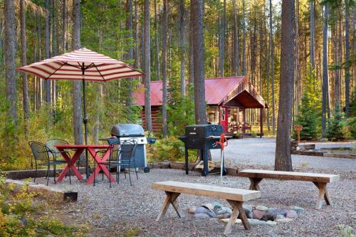 A cozy outdoor campsite with a red table, grill, and shaded seating among tall trees and a cabin in the background.
