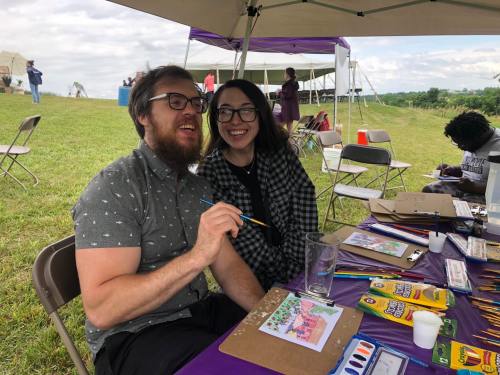 A smiling couple sits at a table with art supplies, enjoying a creative moment outdoors under a tent.