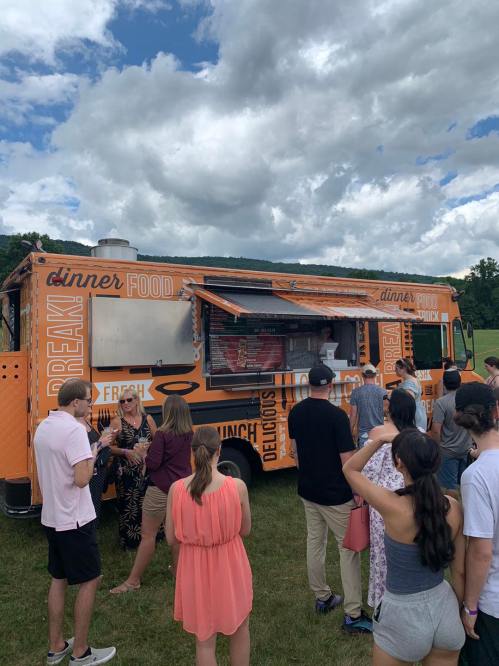 A vibrant food truck with people lined up, enjoying a sunny day with clouds in the background.