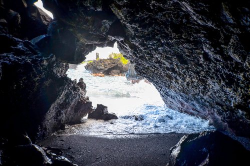 View from inside a rocky cave, looking out at the ocean waves and a rocky shoreline under a bright sky.