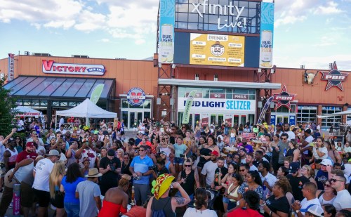 A large crowd gathers at Xfinity Live! for an outdoor event, enjoying drinks and entertainment under a blue sky.