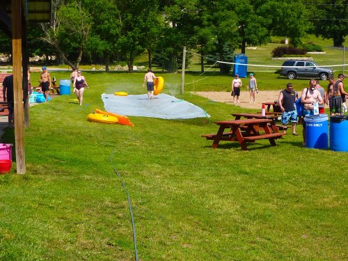 A summer scene with people enjoying a water slide, picnic tables, and a volleyball court in a grassy area.