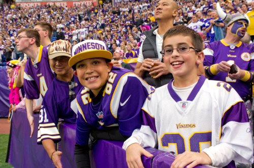 A group of young fans in Minnesota Vikings gear smiles and cheers at a football game, surrounded by a lively crowd.