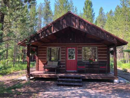 A cozy wooden cabin with a red door, surrounded by tall trees and a gravel path.