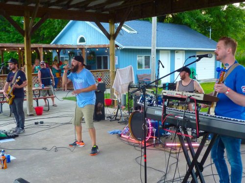 A band performs on stage under a pavilion, with musicians playing instruments and an audience in the background.