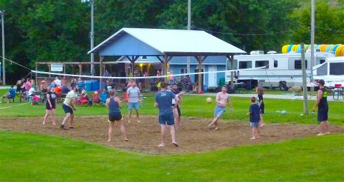 A group of people playing volleyball on a sandy court, with spectators seated nearby and a pavilion in the background.