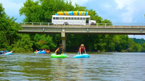 Two people kayak on a river beneath a bridge, with a bus loaded with colorful kayaks parked above.