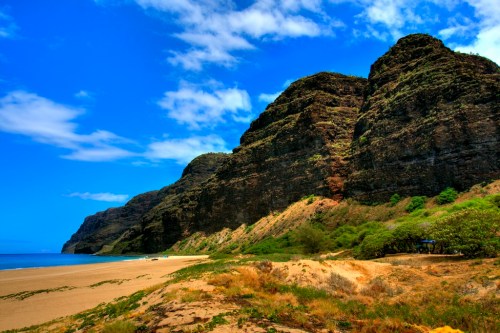 Scenic beach with golden sand, lush greenery, and towering cliffs under a bright blue sky with scattered clouds.