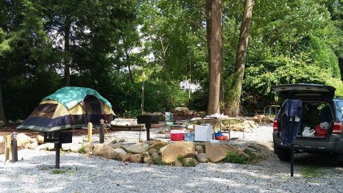 A campsite with a tent, picnic area, and a parked car surrounded by trees and gravel.