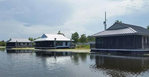 Three modern houses on stilts by a calm water body, surrounded by greenery under a cloudy sky.