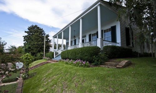 A charming house with a porch, surrounded by greenery and flowers, set on a gentle slope under a blue sky.