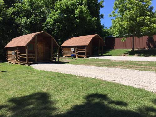 Two wooden cabins sit on a grassy area, surrounded by trees, with a gravel path leading between them.