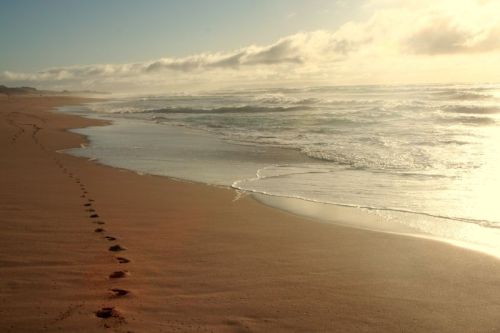 Footprints in the sand lead to a calm beach with gentle waves and a cloudy sky at sunrise.