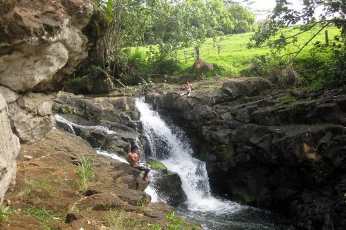 A serene waterfall cascades over rocks, surrounded by lush greenery and two people sitting nearby.