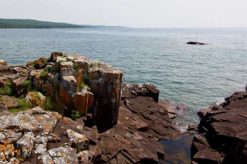 Rocky shoreline with lichen-covered boulders, calm water, and distant hills under a clear sky.