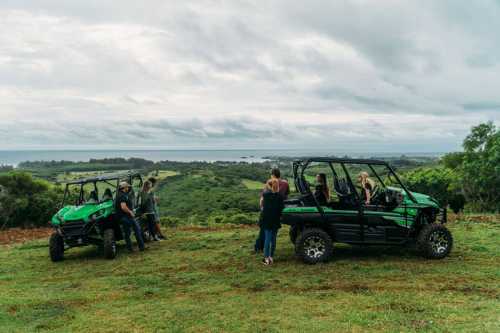 Two green off-road vehicles parked on a grassy hill with a group of people enjoying the scenic view of the landscape.