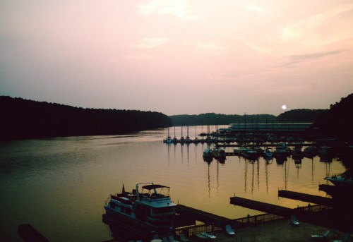 Sunset over a calm lake with boats docked and silhouettes of trees in the background.