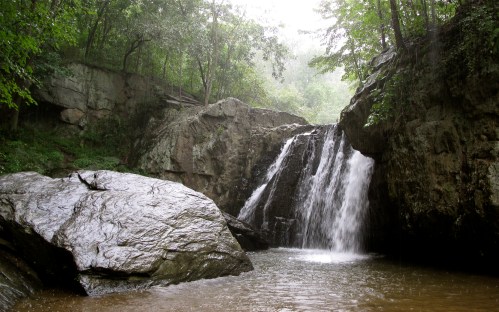 A serene waterfall cascades over rocks into a calm pool, surrounded by lush greenery and misty air.