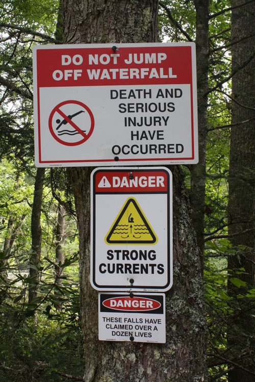 Warning signs on a tree caution against jumping off a waterfall due to strong currents and serious injury risks.