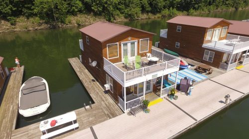 Aerial view of wooden houseboats on a calm lake, with a boat docked nearby and greenery in the background.
