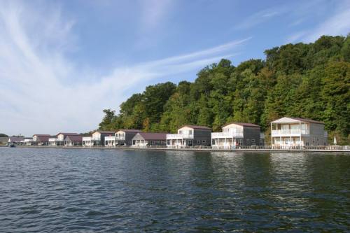 A row of waterfront houses along a calm lake, surrounded by lush green trees under a clear blue sky.