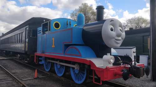 A smiling blue train with a face, parked next to a vintage passenger car on a railway track.