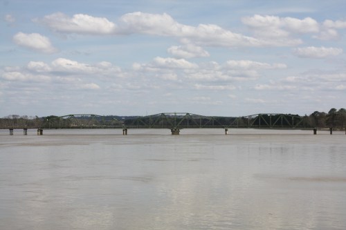 A green bridge spans a calm river under a cloudy sky, with distant hills visible in the background.
