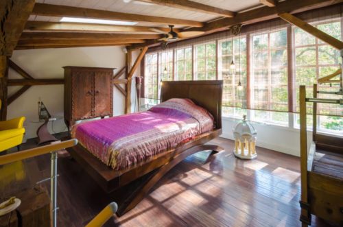 Cozy bedroom with a wooden bed, large windows, and rustic decor, featuring a pink blanket and warm wooden accents.