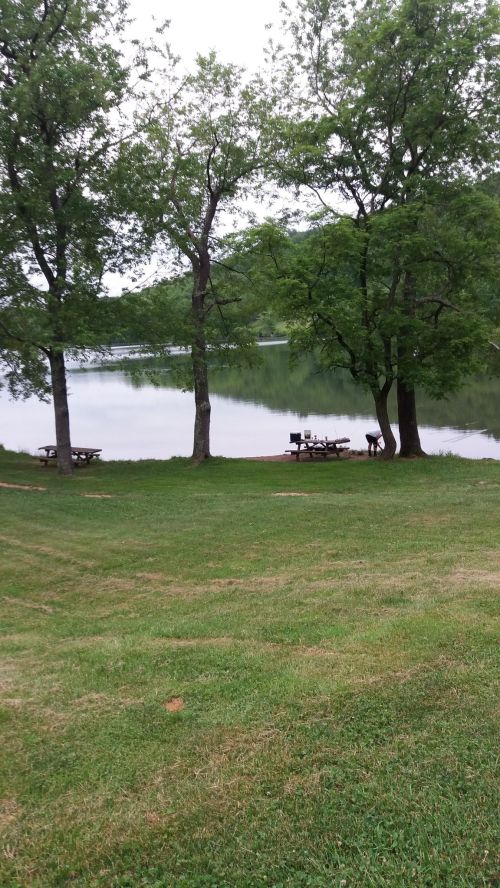 A serene lakeside view with picnic tables under trees, reflecting calm water and green hills in the background.