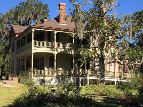 A large, historic two-story house with a wraparound porch, surrounded by trees and Spanish moss.