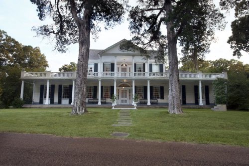 A large, two-story white house with a porch, surrounded by trees and a grassy lawn.