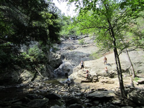 A rocky landscape with people exploring near a stream, surrounded by trees and steep slopes.