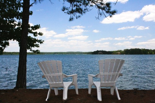 Two white chairs facing a serene lake under a blue sky with fluffy clouds, surrounded by trees.