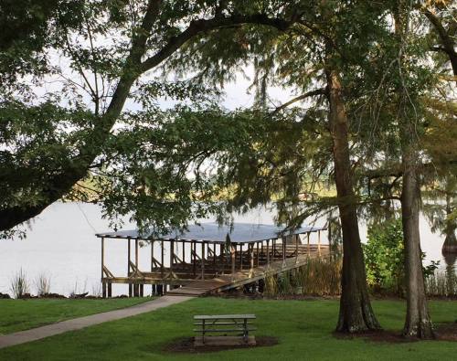 A wooden dock extends into a calm lake, surrounded by trees and a grassy area with a picnic table.