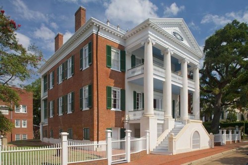 A historic brick building with white columns, green shutters, and a white fence, surrounded by trees and blue sky.