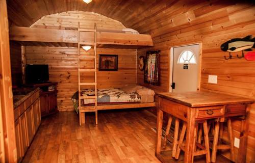 Cozy wooden cabin interior with a lofted bed, kitchenette, and a small dining table. Warm, rustic decor.